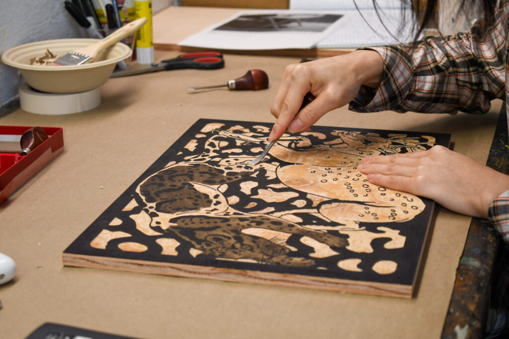 Close-up of Jenie Gao’s hands carving a woodblock of a Formosan Sika Deer and Clouded Leopard in a cyclical chase.